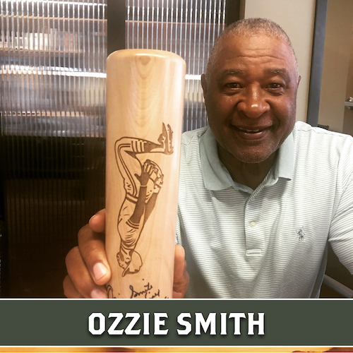 Ozzie Smith smiling and holding up a wooden Dugout Mugs baseball bat mug engraved with a portrait.